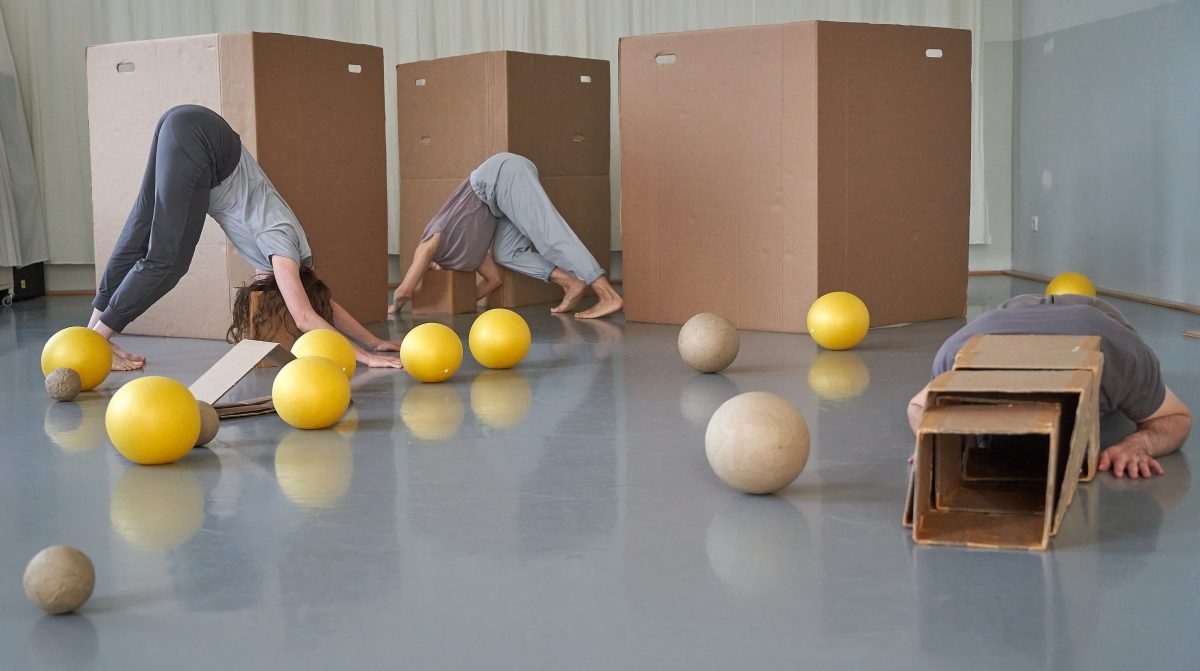 Zwei Tänzerinnen und ein Tänzer auf einer Bühne. Sie stecken mit den Köpfen in Quadraten fest, d.h. eigentlich in quadatischen Kartons. Auf der Bühne liegen goldene und hellbraune Kugeln in verschiedenen Größen.