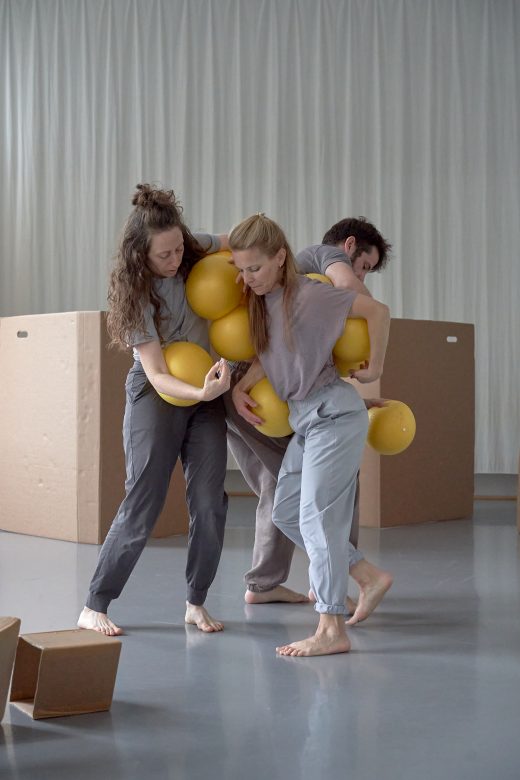 Zwei Tänzerinnen und ein Tänzer auf einer Bühne. Sie versuchen gemeinsam, acht goldfarbene Kugeln festzuhalten. Auf der Bühne liegen Kartons, die verschiedene geometrischen Figuren darstellen.