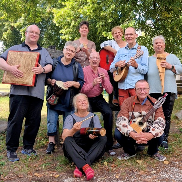 Neun freundliche Menschen sitzen und stehen zusammen auf einer Wiese in einem Park. Es ist Sommer und alle haben ein Instrument in der Hand, z. B. eine Gitarre und eine Panflöte.