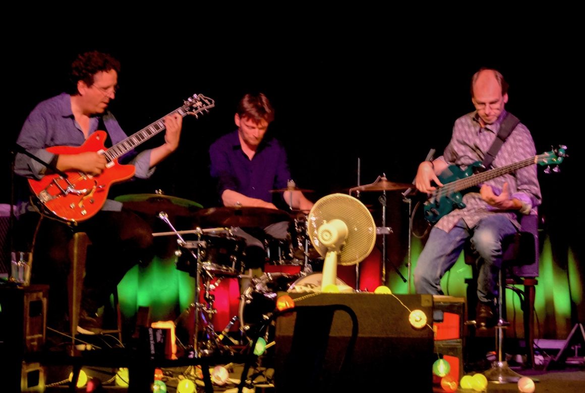 Drei Jazzmusiker spielen auf einer Bühne: Schlagzeug, Gitarre und E-Bass.