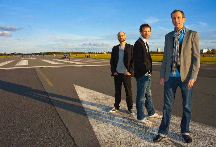 Die drei Musiker des Lionel Haas Trio stehen auf dem Tempelhofer Feld in Berlin unter blauem Himmel.