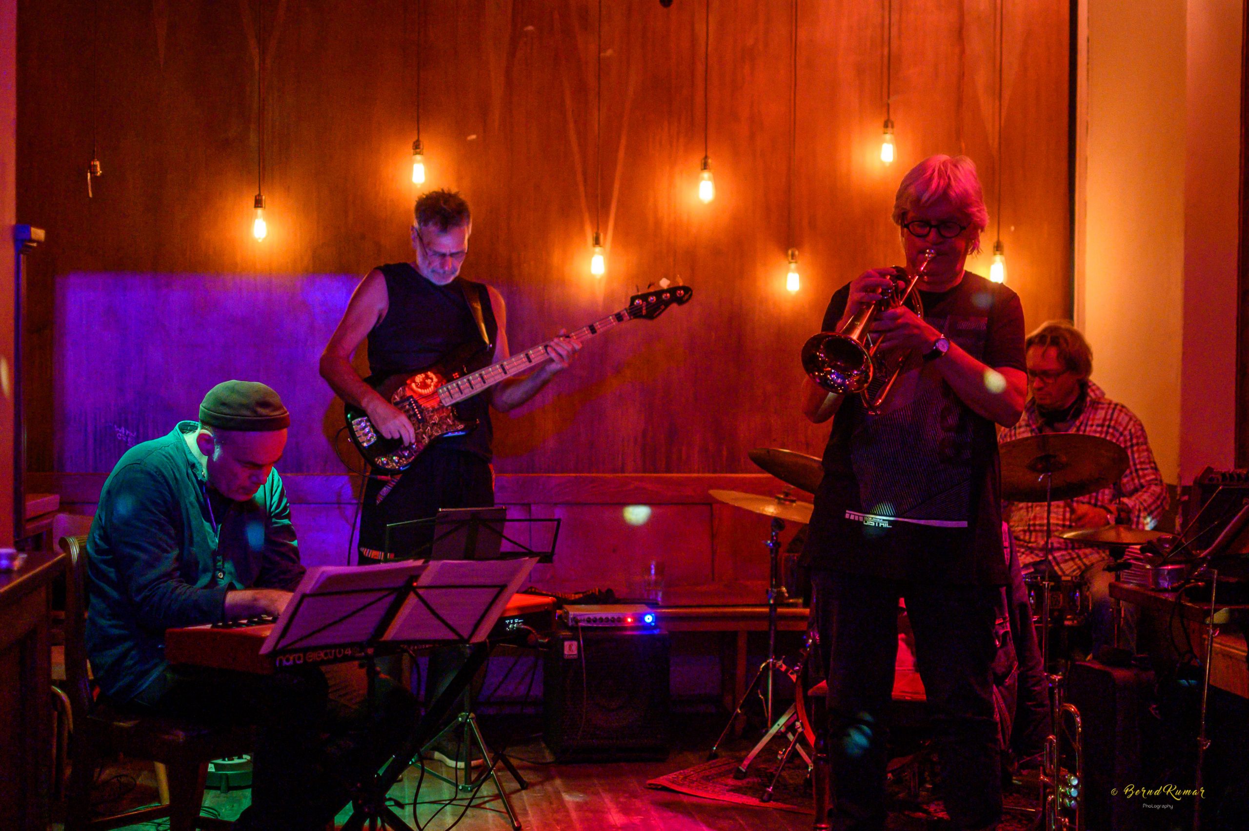 drei Musiker auf der Bühne. Im Hintergrund sind Lampen