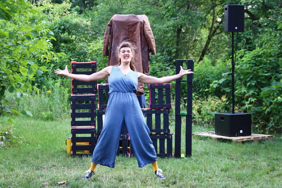 Eine Schauspielerin steht mit ausgebreiteten Armen und wütendem Gesicht in einem Garten