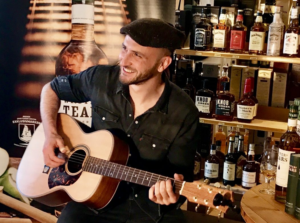 Mann mit Gitarre, im Hintergrund ein Regal mit Flaschen