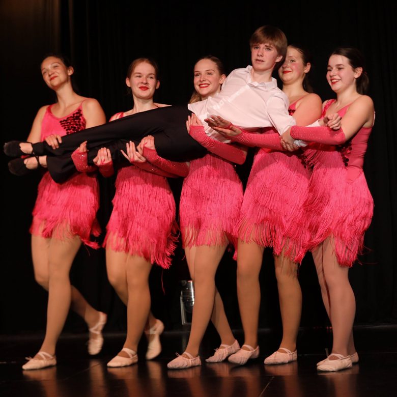 Fünf junge Frauen mit flamingofarbigem Revue-Dress halten mit ihren Armen einen jungen Mann hoch