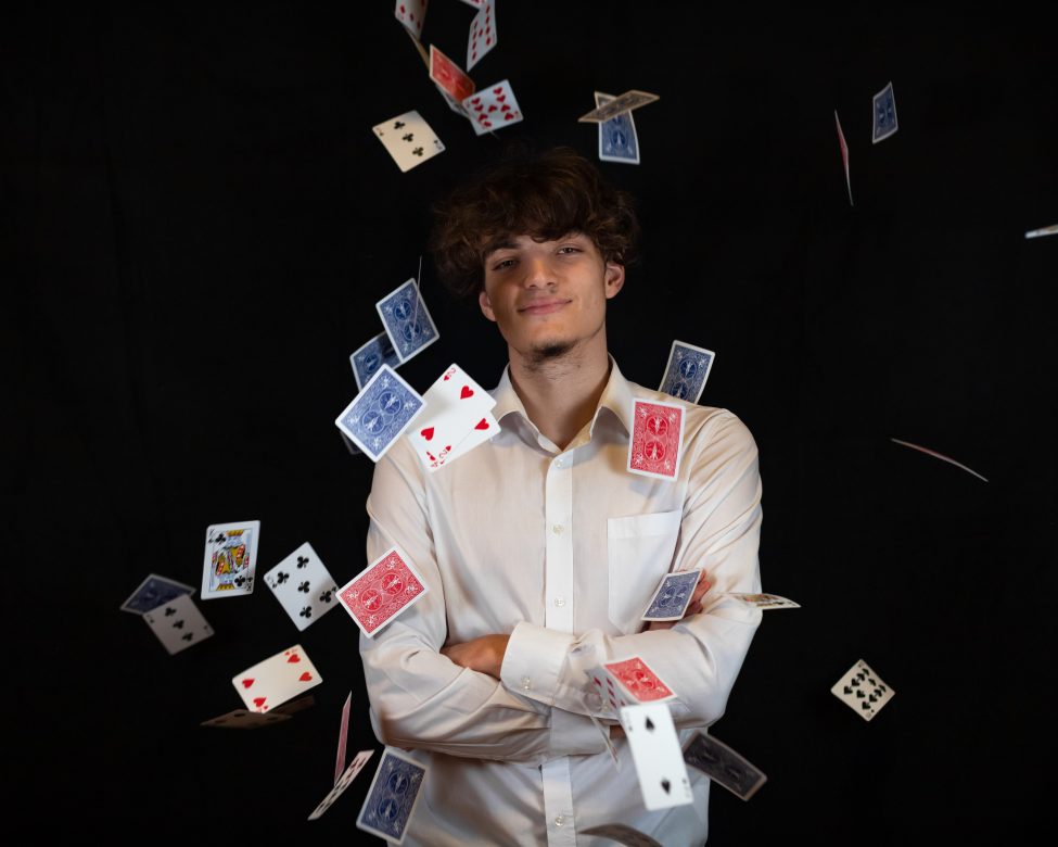 Der Zauberkünstler Ben-Arik Mann sieht lächelnd in die Kamera, um ihn herum schweben zahlreiche Spielkarten