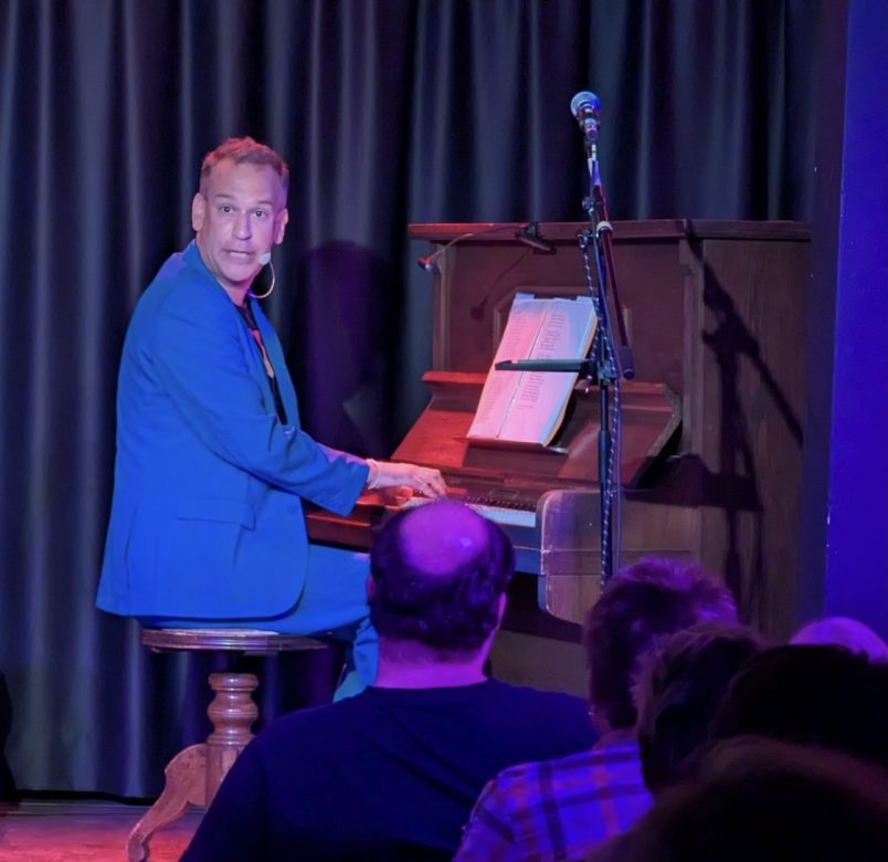 Der Kabarettist Tom Kaminsky spielt am Piano auf einer Bühne und singt, wobei er ins Publikum blickt.