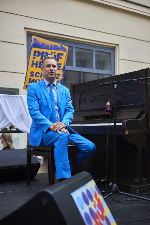 Der Kabarettist Tom Kaminsky sitzt in einem hellblauen Anzug auf einer Bühne im Freien an einem Piano. Er sieht freundlich in die Kamera.